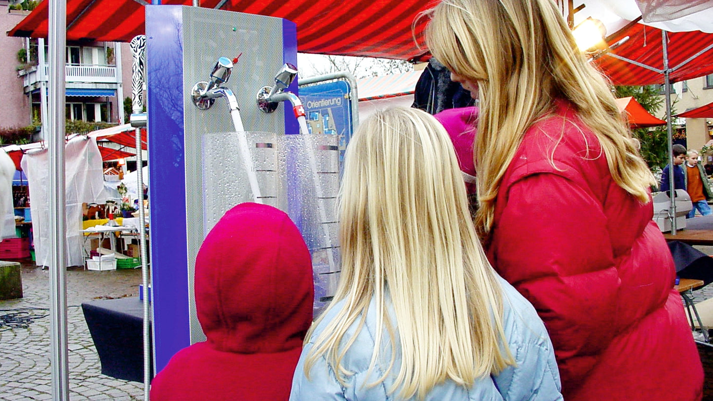 Sur une place de marché, une démonstratrice montre le fonctionnement d'AquaClic sur une fontaine de démonstration.